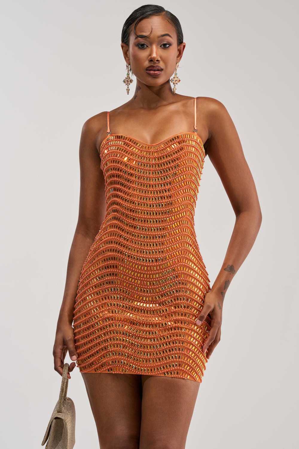 Dressed in the SHINE ON RHINESTONE MINI DRESS IN ORANGE—a shimmering, sequin-adorned mini with slender straps—she stands confidently, clutching a small beige handbag. With her slicked-back hair and dangling earrings, she embodies the ultimate show-stopping look for anyone searching for a stunning bodycon birthday dress.
