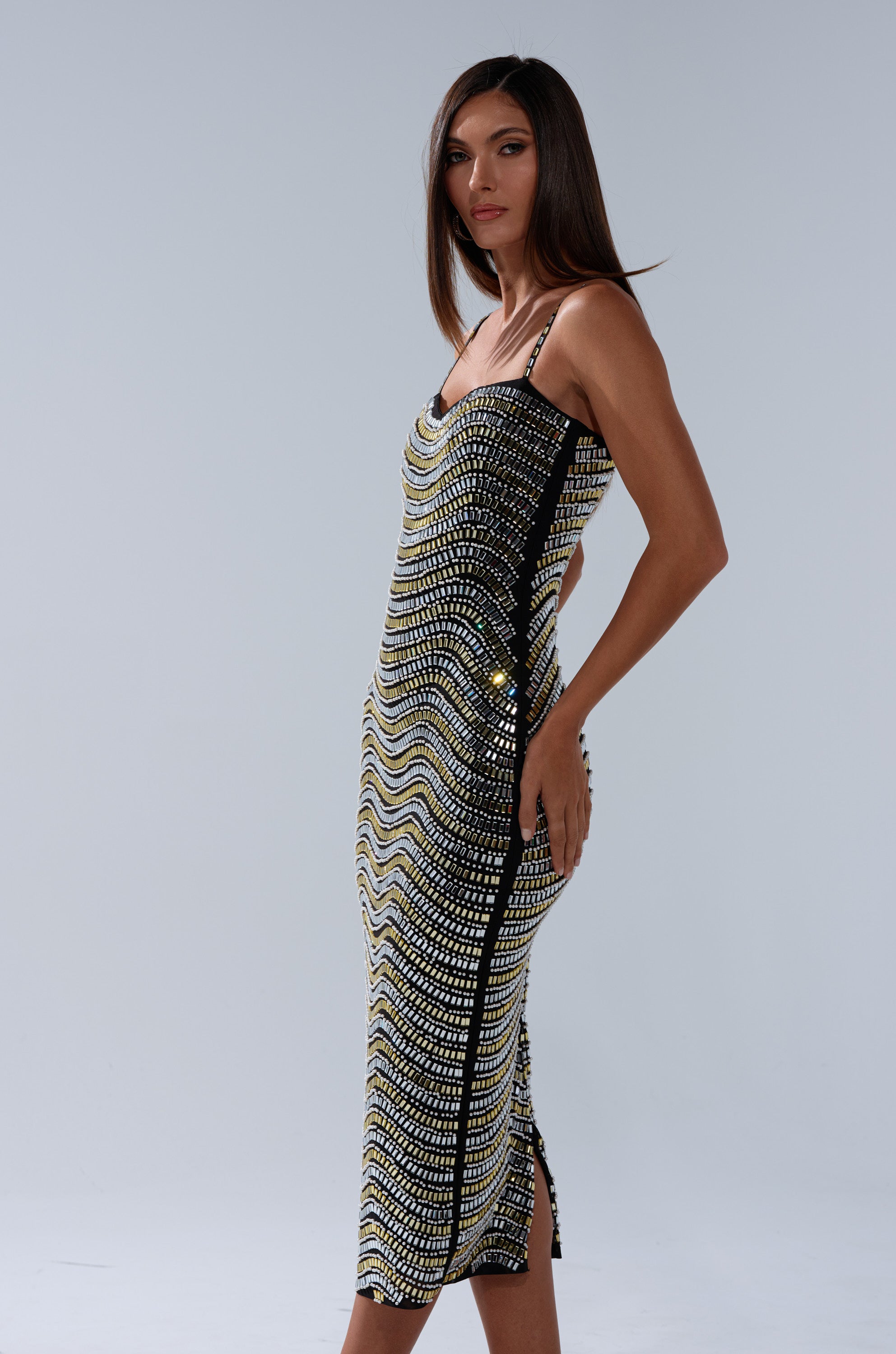 A woman poses against a plain grey background in the SHINE ON RHINESTONE MIDI DRESS, featuring a curve-hugging fit and black, gold, and white sequin wavy pattern. She has straight brown hair and rests one hand on her hip.