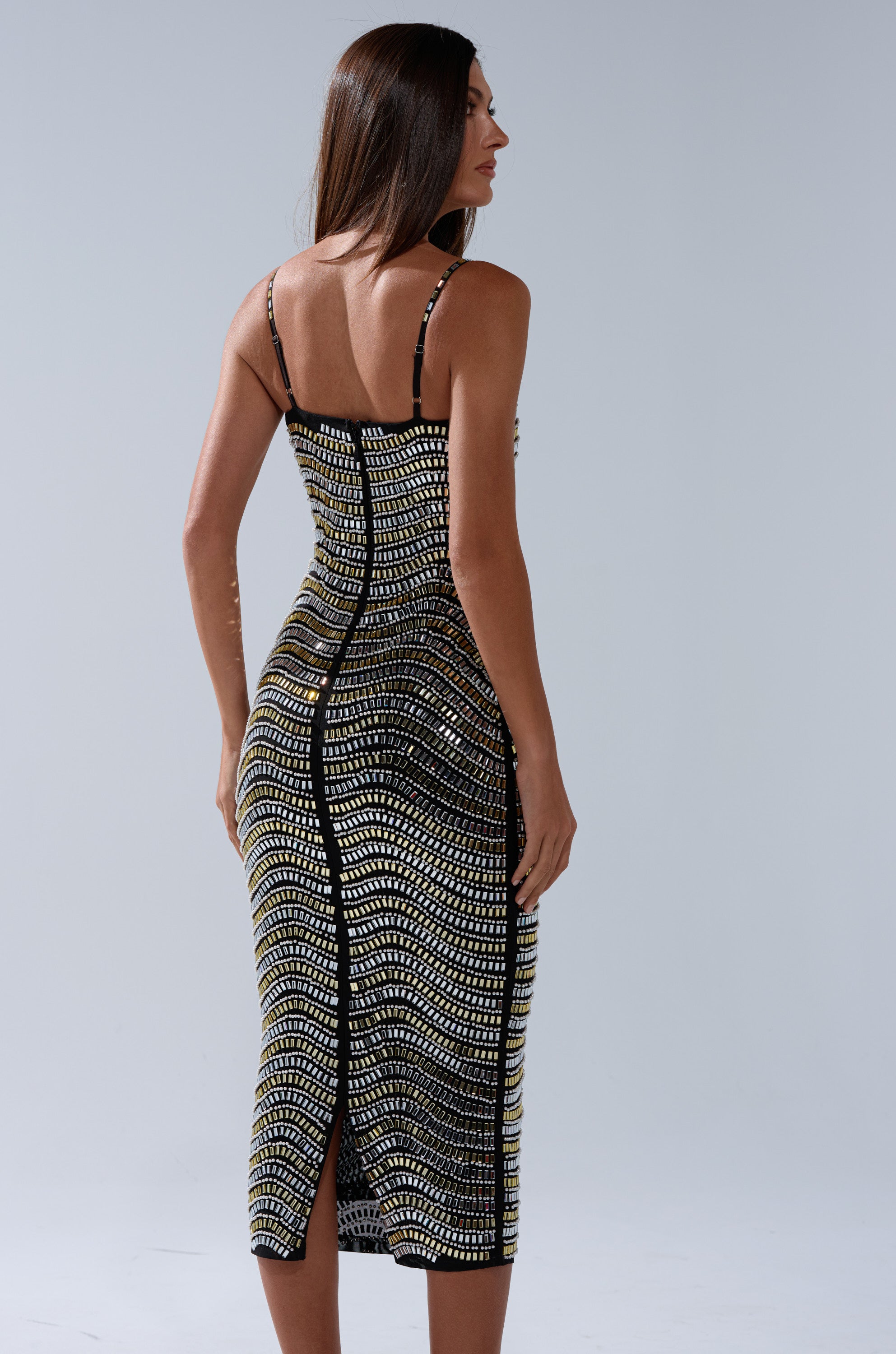 Woman with long brown hair wearing the SHINE ON RHINESTONE MIDI DRESS in black and gold, viewed from the back, standing against a plain light gray background.