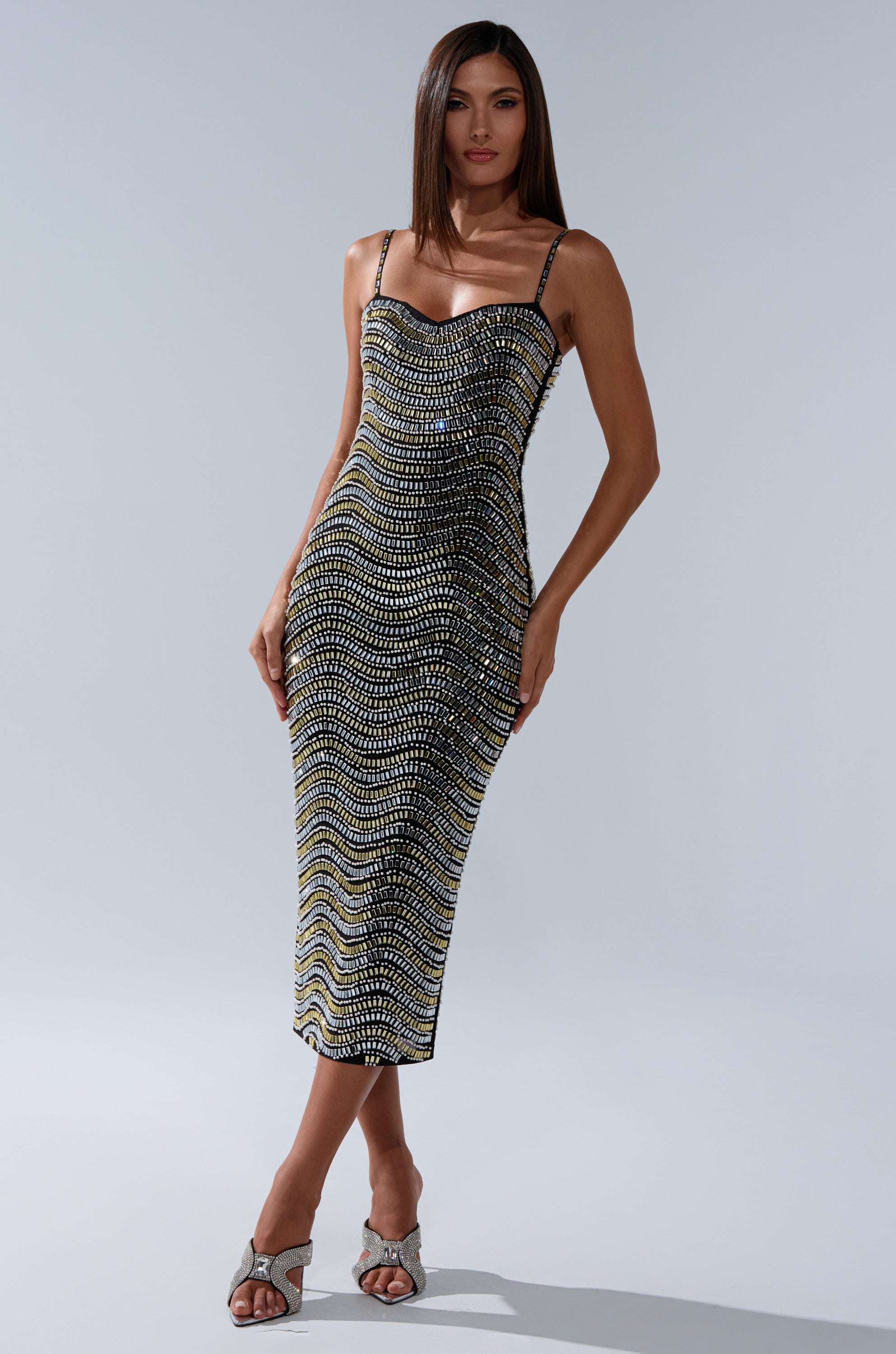 A woman with long brown hair and a neutral expression wears the SHINE ON RHINESTONE MIDI DRESS, a form-fitting mesh mid-length dress with wavy black, white, and yellow patterns, paired with open-toed heels against a plain backdrop.