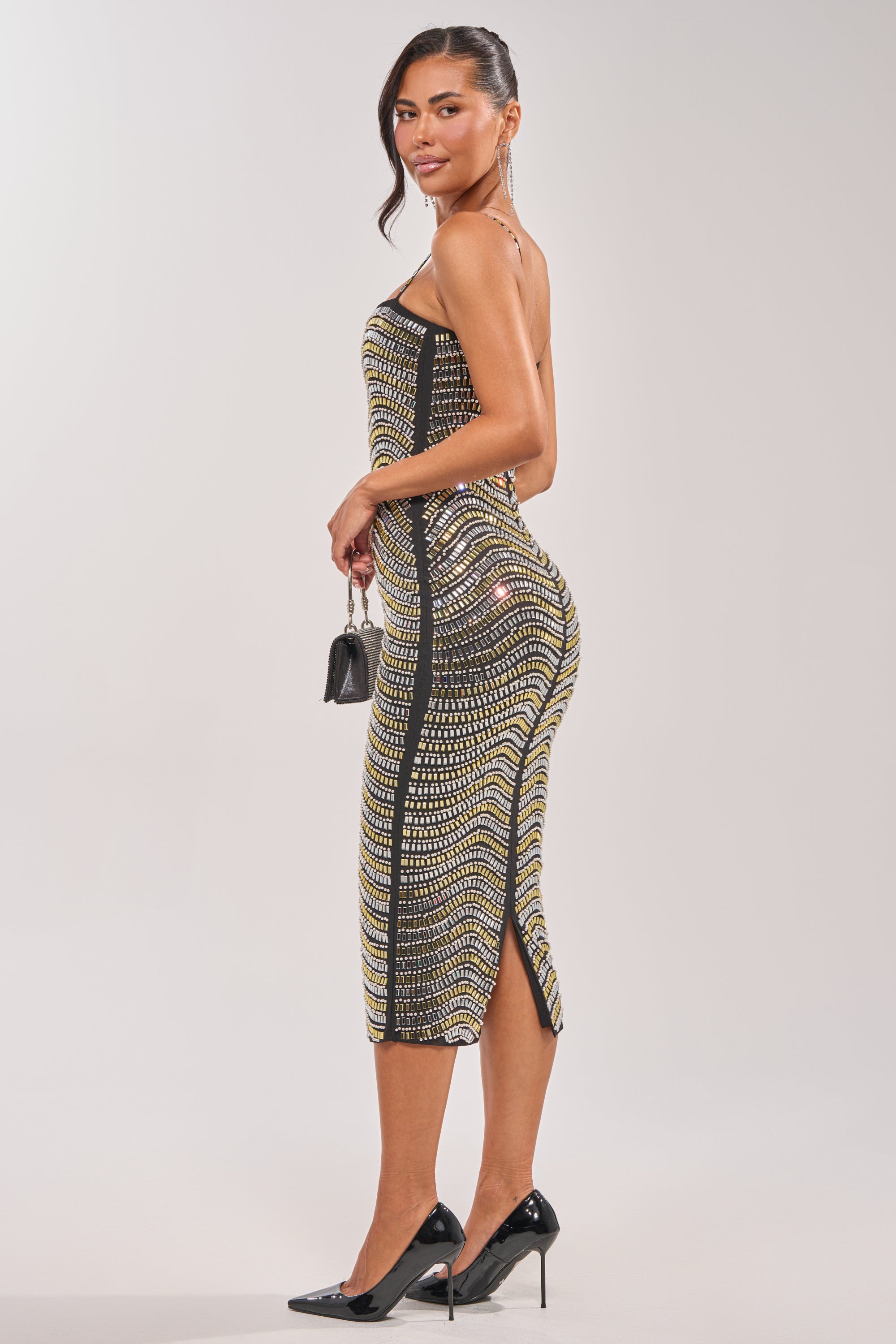 A woman stands against a plain light background, looking over her shoulder in the SHINE ON RHINESTONE MIDI DRESS with a black and yellow pattern, side slit, black heels, and a small black handbag. 5