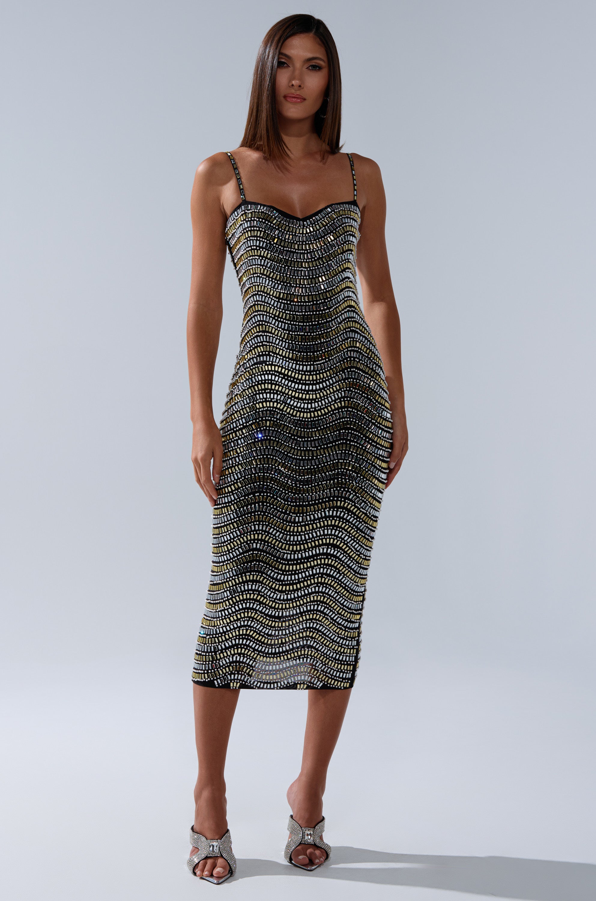 A woman poses in a studio wearing the SHINE ON RHINESTONE MIDI DRESS, a fitted, sleeveless mesh dress with black and gold wavy patterns. She pairs it with silver open-toe heels, and her straight brown hair stands out against the light gray background.
