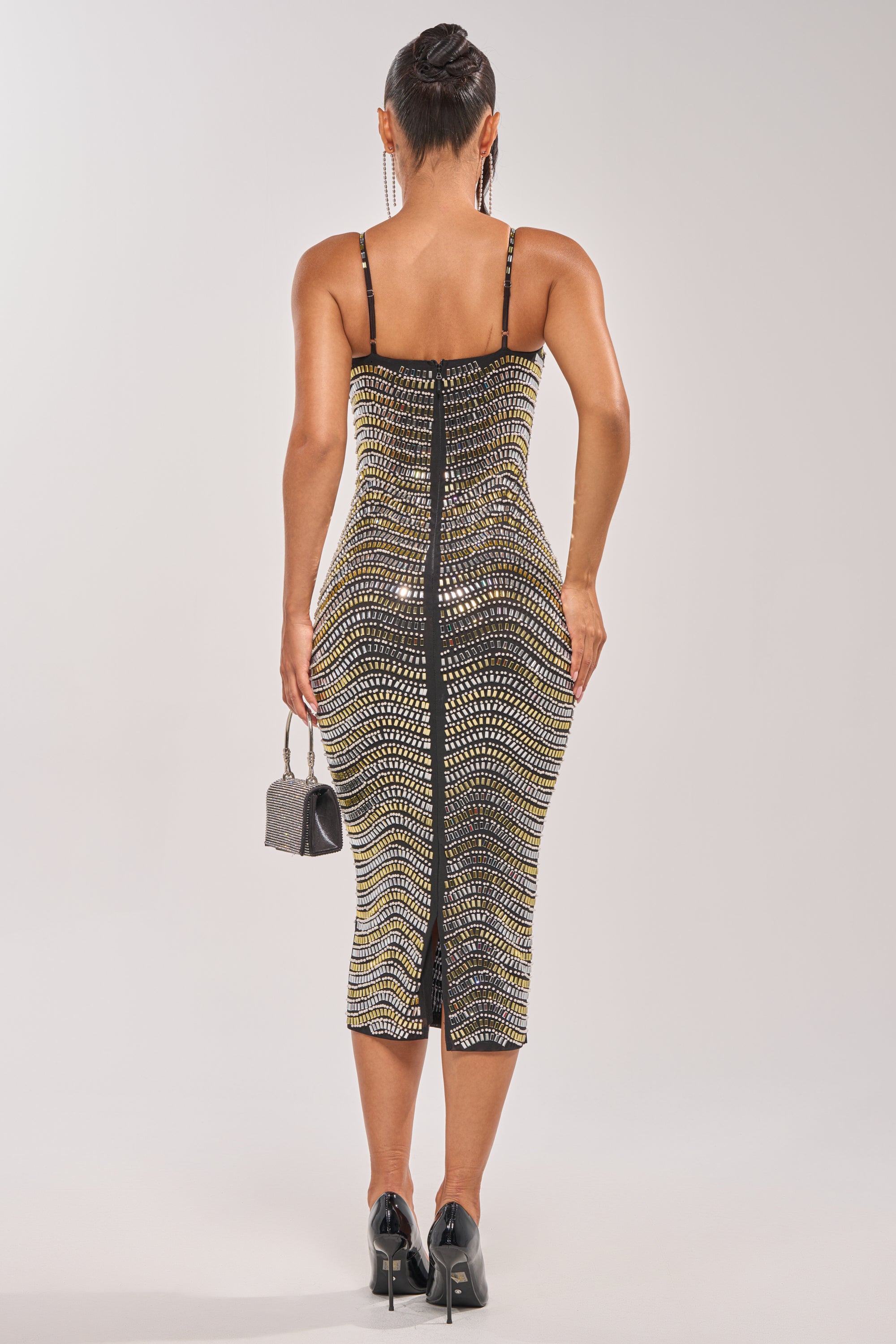 A woman faces away, wearing the SHINE ON RHINESTONE MIDI DRESS with black and gold sequins, spaghetti straps, and a back zipper. She holds a glittery handbag and pairs the dress with black high heels. 6