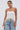 Woman wearing the PEARL TRIM HIGH LOW CORSET with decorative fringe and light blue high-waisted jeans, holding a white woven clutch, poses against a plain light background.