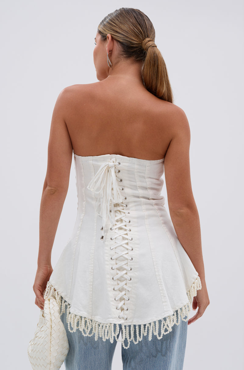 A woman with long, straight hair in a low ponytail, facing away, wears the PEARL TRIM HIGH LOW CORSET featuring a strapless design, lace-up back, and decorative trim. She pairs it with light blue jeans and holds a small white bag.