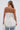 A woman with long, straight hair in a low ponytail, facing away, wears the PEARL TRIM HIGH LOW CORSET featuring a strapless design, lace-up back, and decorative trim. She pairs it with light blue jeans and holds a small white bag.