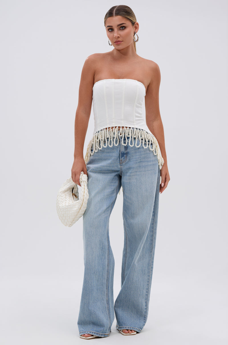 A woman stands facing forward, wearing the PEARL TRIM HIGH LOW CORSET, wide-leg blue jeans, and heeled sandals. She holds a woven white clutch, has her hair pulled back, and accessorizes with large hoop earrings.