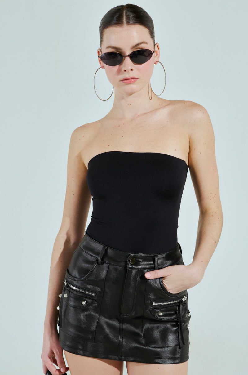 Wearing THE ULTIMATE TUBE PAXTON BODYSUIT in black, a person cinches their curves effortlessly against a light blue backdrop. Large hoop earrings and trendy sunglasses complete the look, while one hand is tucked into their black leather mini skirt's pocket.