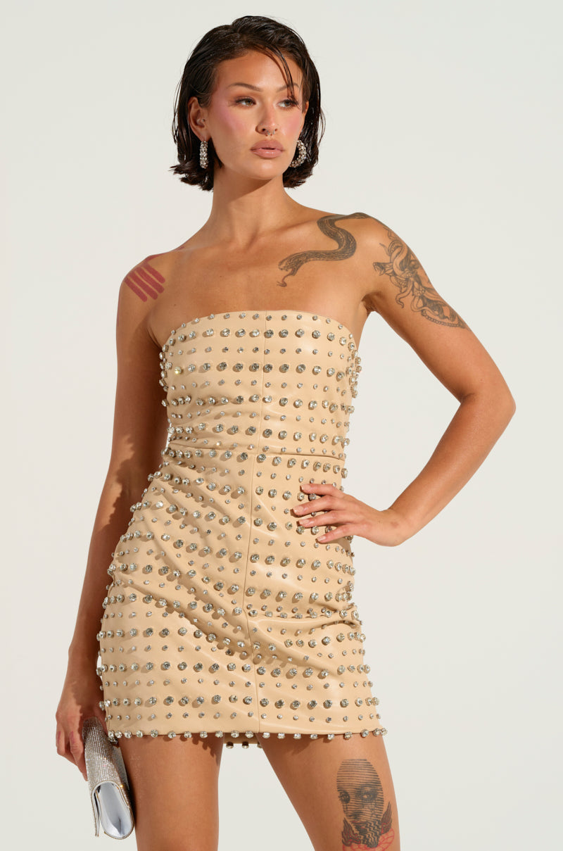 A person with short dark hair stands confidently in the TELL ME ABOUT IT STUD PU MINI DRESS, a strapless beige faux leather dress featuring metal studs. They carry a silver clutch decorated with rhinestones and showcase several tattoos on their arm and shoulder.