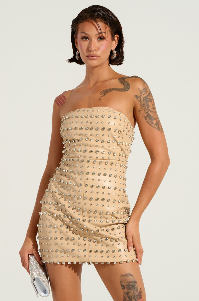 A person with tattoos confidently poses in the TELL ME ABOUT IT STUD PU MINI DRESS, a strapless beige faux leather creation glistening with silver rhinestone embellishments. They hold a small silver purse, and the plain background highlights the edgy outfit and bold stance.