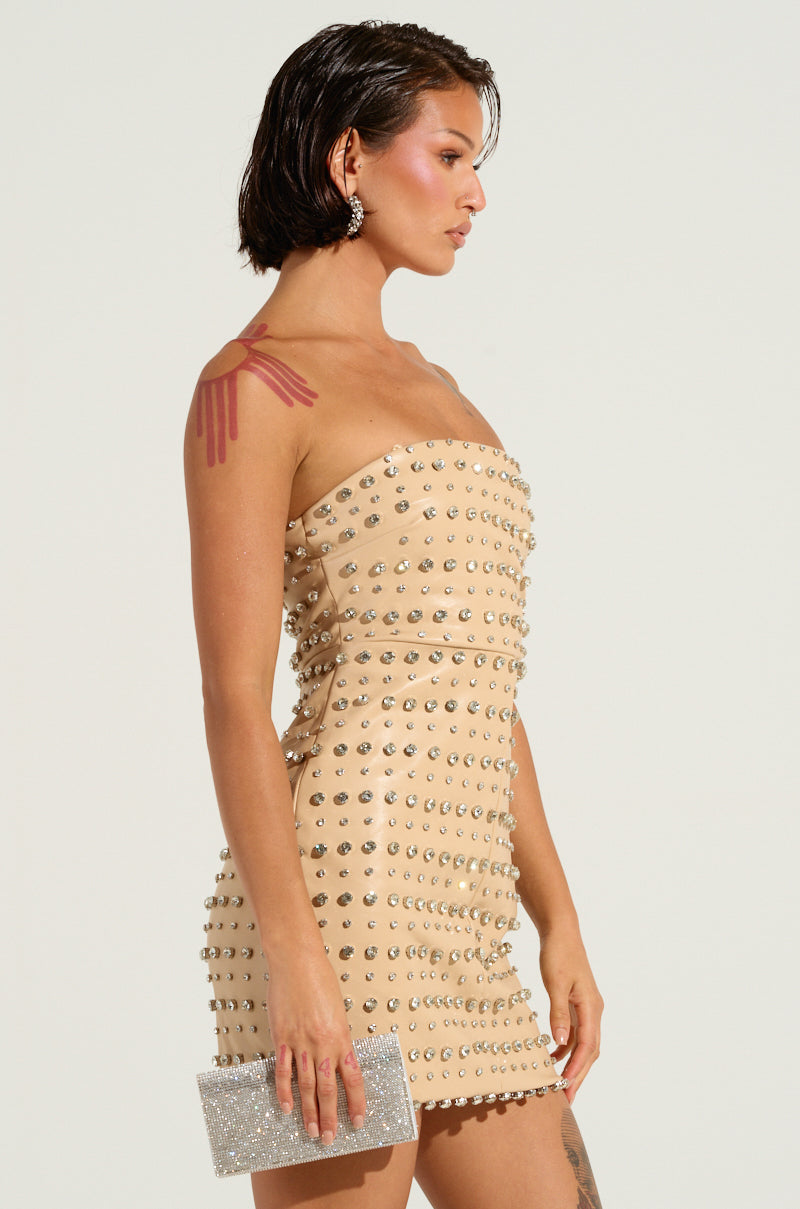 A woman with wet-look hair poses in the TELL ME ABOUT IT STUD PU MINI DRESS, a strapless beige outfit adorned with silver rhinestone embellishments. She holds a glittery silver clutch and wears stud earrings. A tattoo is visible on her shoulder and arm against the plain light gray background.