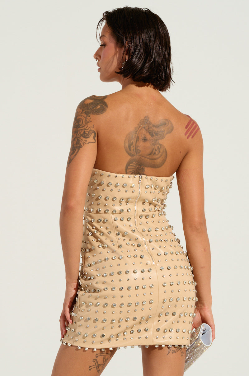 Facing away from the camera, a person with short hair is wearing the TELL ME ABOUT IT STUD PU MINI DRESS, featuring silver rhinestone embellishments. Their arms and back are covered in various tattoos as they hold a small clutch in their right hand against a simple, light-colored background.