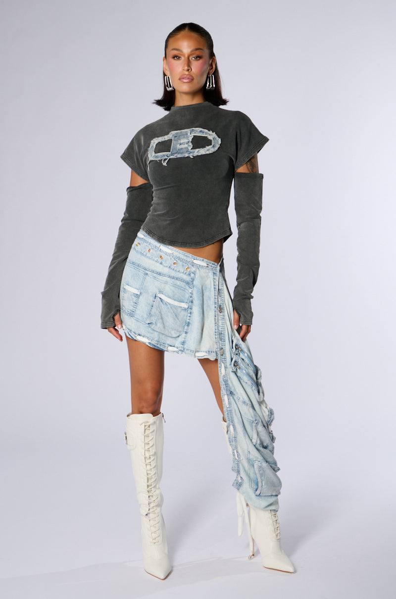 A woman stands against a plain backdrop wearing a distressed gray crop top, the Chambray Denim Wrap Around Mini Skirt featuring an acid wash finish and hanging fabric on one side, and white knee-high lace-up boots. She accessorizes with geometric earrings and wears long dark gray fingerless gloves.