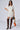 A woman stands posing in a white Kirby Ruched Tshirt Mini Dress with cut-out and gathering detail on one side. She pairs it with brown, over-the-knee boots featuring patterned embellishments and holds a black handbag adorned with an orange and white patterned scarf.