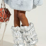 A person wearing a fluffy light blue coat and the OUTTA THIS WORLD PUFFER LEGWARMERS in an oversized metallic design stands with legs crossed. Their feet are encased in pointed silver shoes, and they hold a red textured purse with adjustable bungee cords against a light, neutral background.