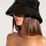 A woman with long brown hair, partially covering her eyes with the TIME TO SHINE SEQUIN BUCKET HAT IN BLACK, is dressed in a strapless, colorful top featuring abstract patterns by the AKIRA Label. She stands against a plain light background.