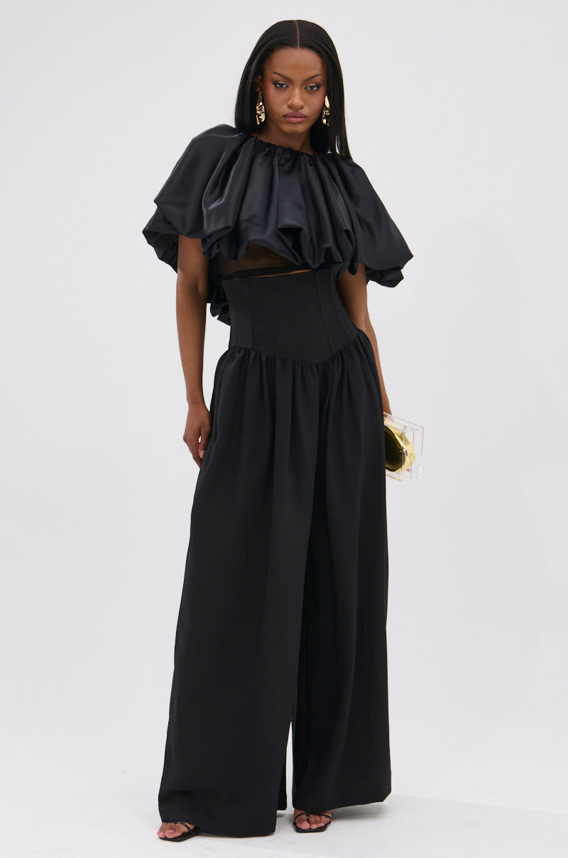A woman in a dramatic black outfit with a ruffled cropped top, statement earrings, clear box clutch, and BETTER THAN THIS CORSETED TROUSER—featuring a high-rise waistband and wide-leg silhouette—stands against a plain white background.