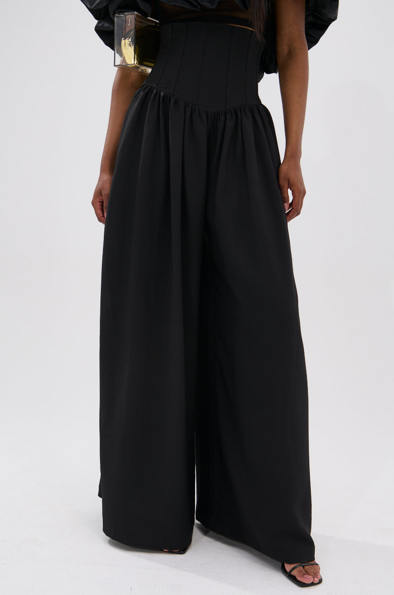 A person wears the BETTER THAN THIS CORSETED TROUSER—wide-leg black pants with a high-rise waist—paired with black open-toe shoes, standing against a plain light background. Only the lower body and part of one arm are shown.