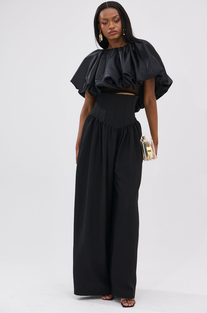 A woman poses against a white background wearing a black ruffle top, gold earrings, black sandals, and the BETTER THAN THIS CORSETED TROUSER—high-waisted wide-leg pants—holding a small clear clutch and sporting straight hair.