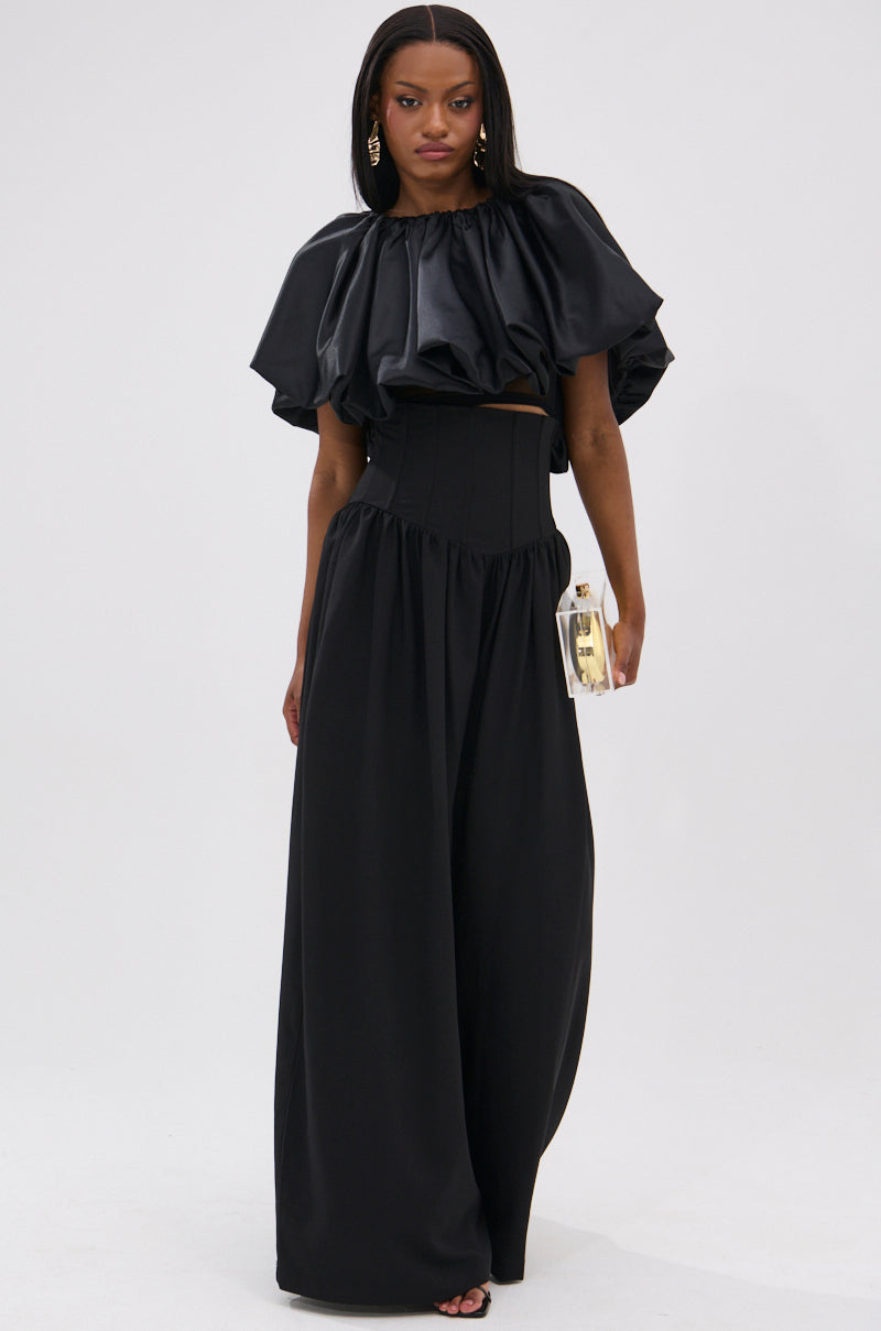 A woman stands against a white background wearing high-waisted, wide-leg BETTER THAN THIS CORSETED TROUSER pants with a black ruffled top, holding a metallic clutch and accessorized with statement earrings.