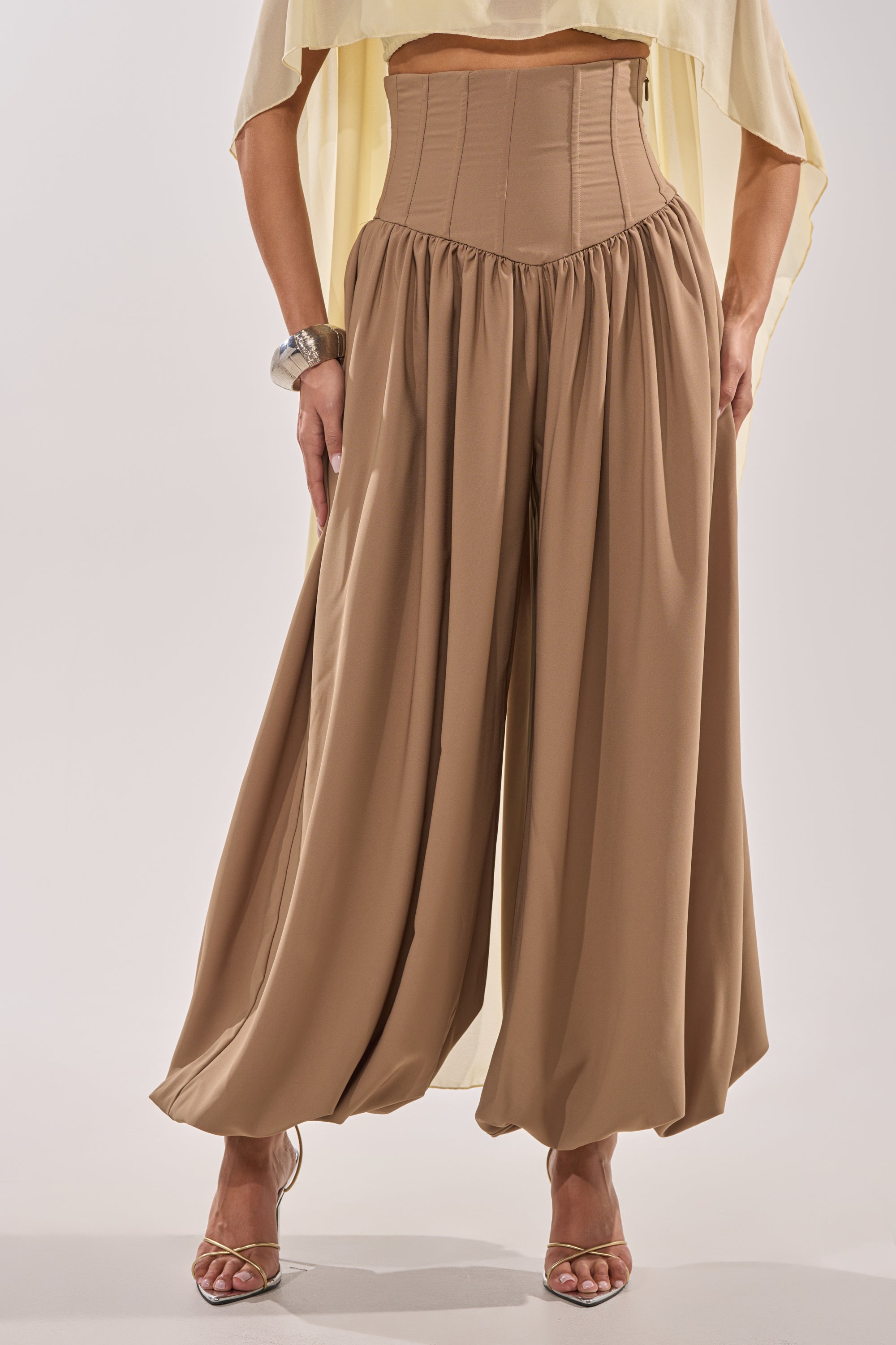 A person wearing FEELING LUXE CORSETED BALLOON PANT in tan, featuring a high-rise waistband and corset-like details, styled with a flowy yellow top, metallic bracelet, and strappy heeled sandals. Face is not visible.