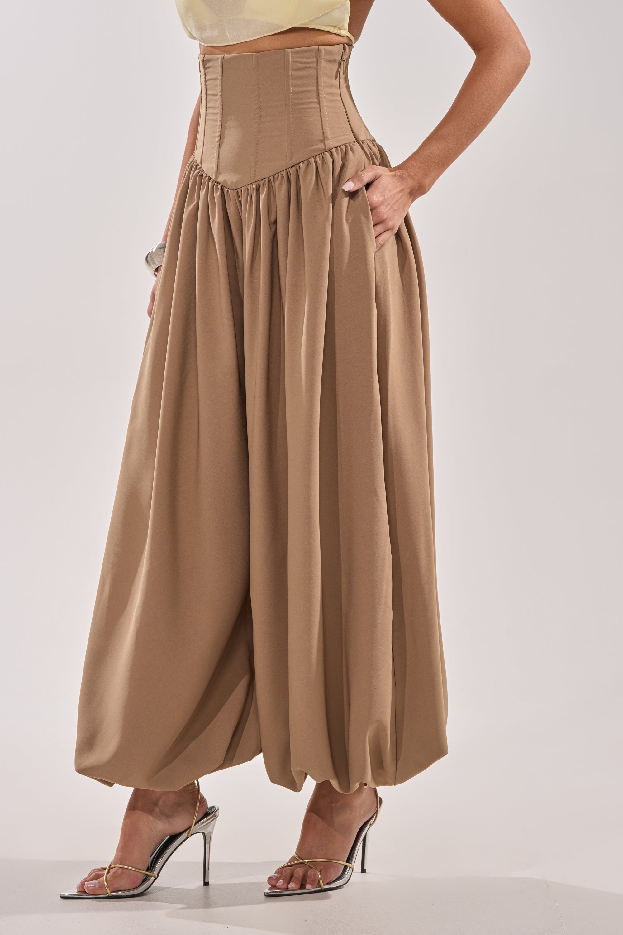 A person wears the FEELING LUXE CORSETED BALLOON PANT—tan, high-rise, loose-fitting pants with a gathered waist and voluminous silhouette—styled with heeled sandals for a chic look. 1