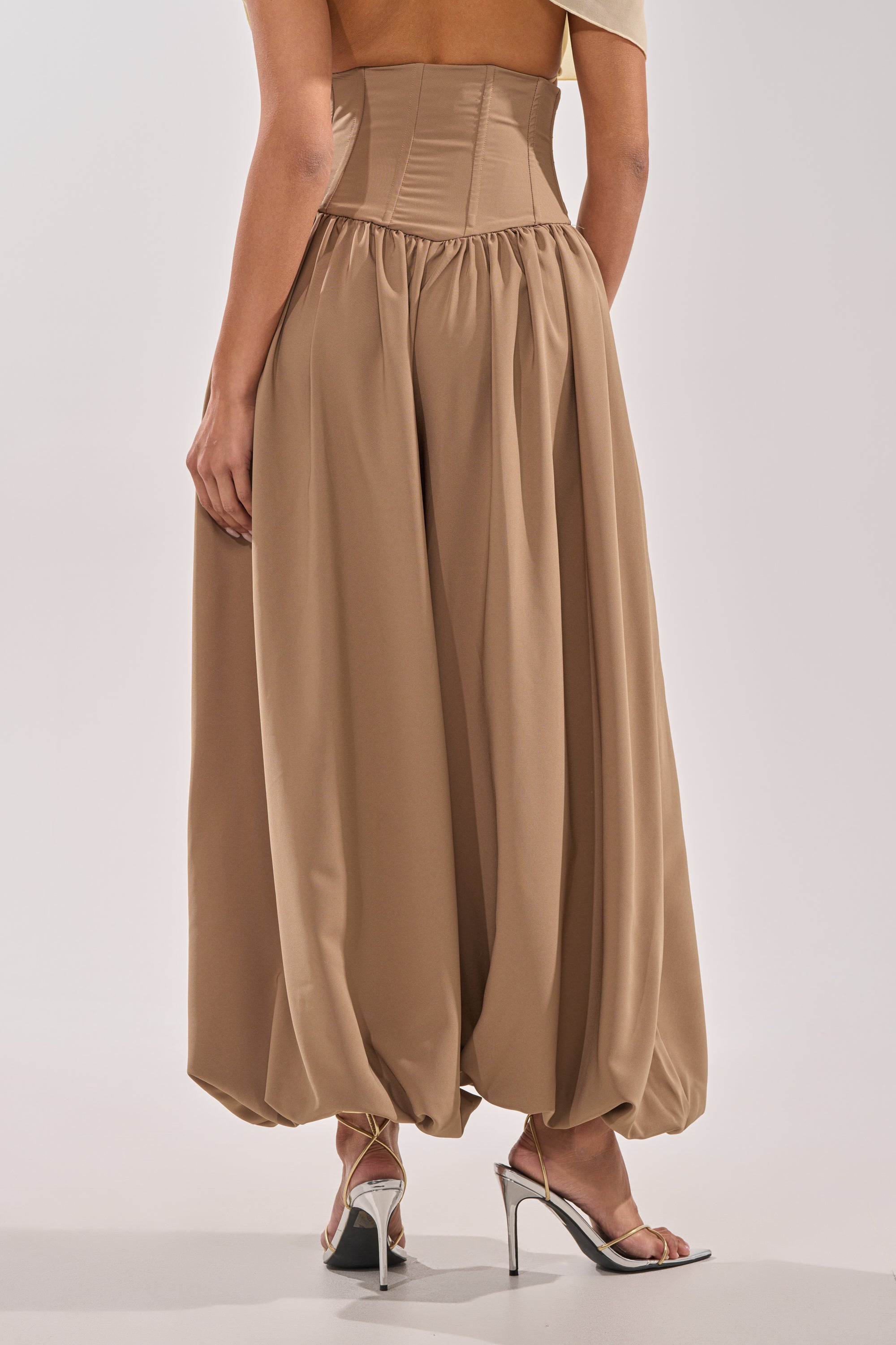 Someone wears the FEELING LUXE CORSETED BALLOON PANT in tan, featuring a flowy, high-rise waistband design, paired with silver high-heeled sandals against a plain light background. 2