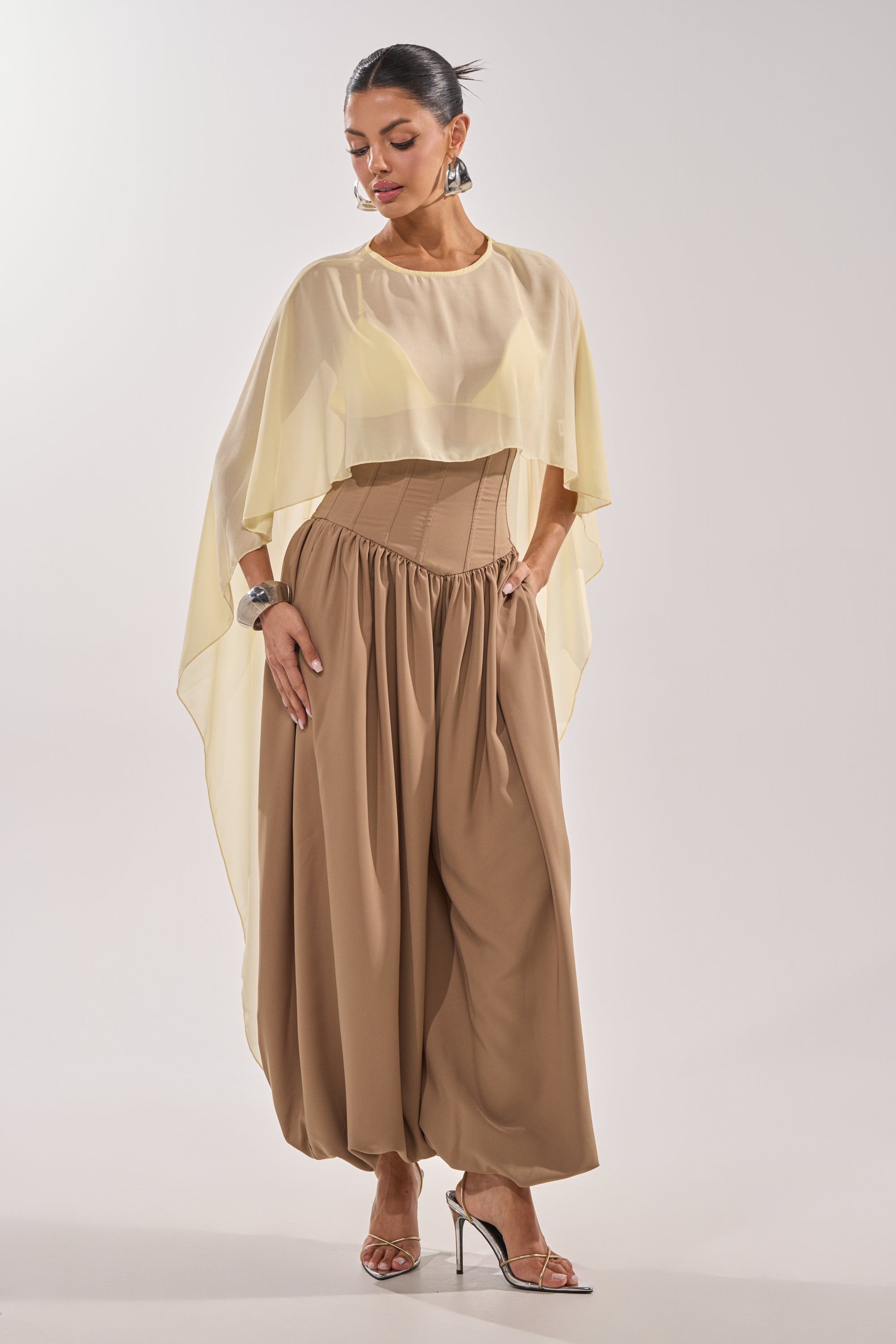 A woman models the FEELING LUXE CORSETED BALLOON PANT in taupe, styled with a beige corset belt, sheer pale yellow cape top, hoop earrings, bracelet, and silver heels against a plain white background. 3