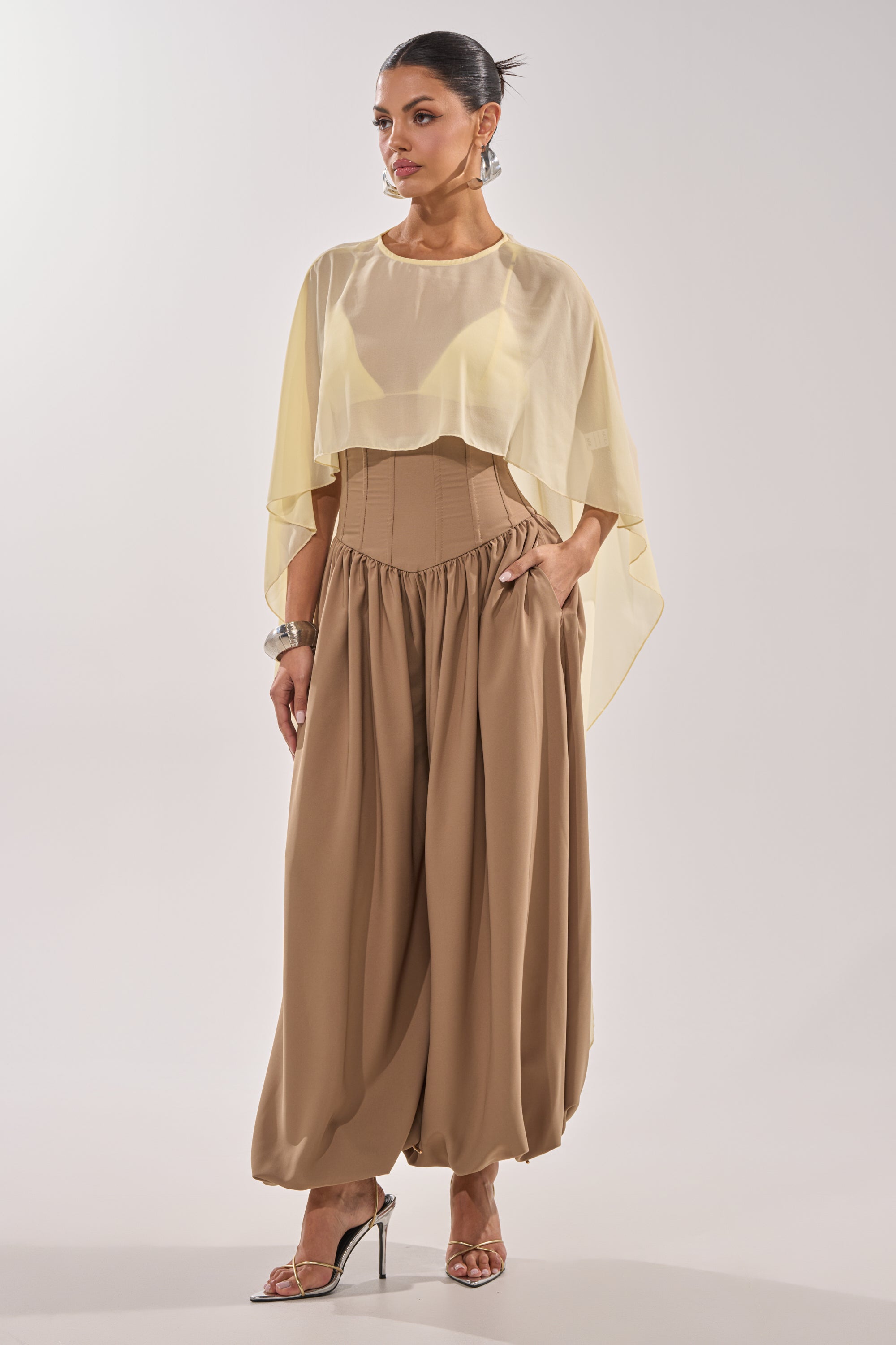 A woman models the FEELING LUXE CORSETED BALLOON PANT, styled with a sheer light yellow cape top, sleek bun, metallic heels, and silver bangles. 4