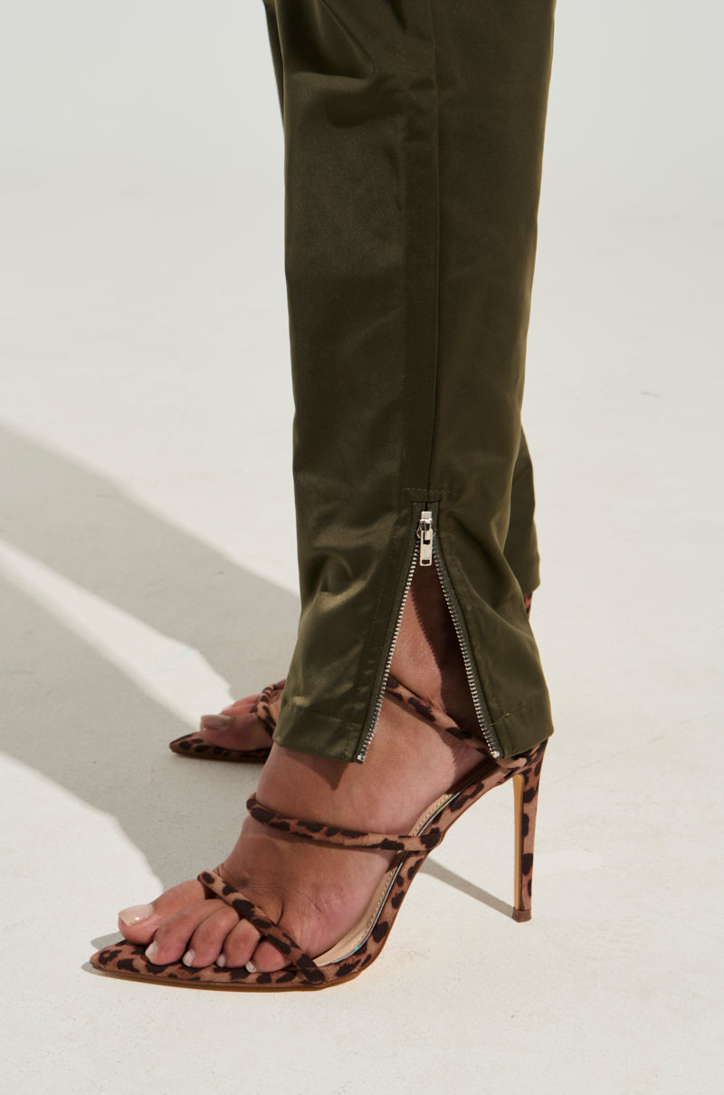 A person wearing the NOT MY PROBLEM TROUSER, a pair of high-rise straight-leg pants with zippered cuffs, paired with leopard-print high-heeled sandals. The pants are unzipped at the bottom, revealing part of the person's ankle and foot. The background is a plain, light-colored surface.