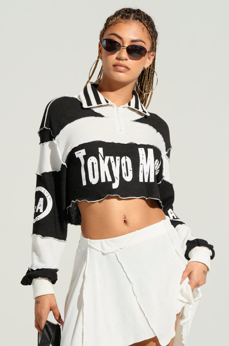 A person with braided hair is wearing black sunglasses and a black and white cropped sweater from the "HANDMADE AND COOL TOP" collection, featuring text that reads "Tokyo MOTO." They pair it with a white asymmetrical skirt. The outfit's chic colorblock paneled construction stands out as they pose against a plain, light-colored background.