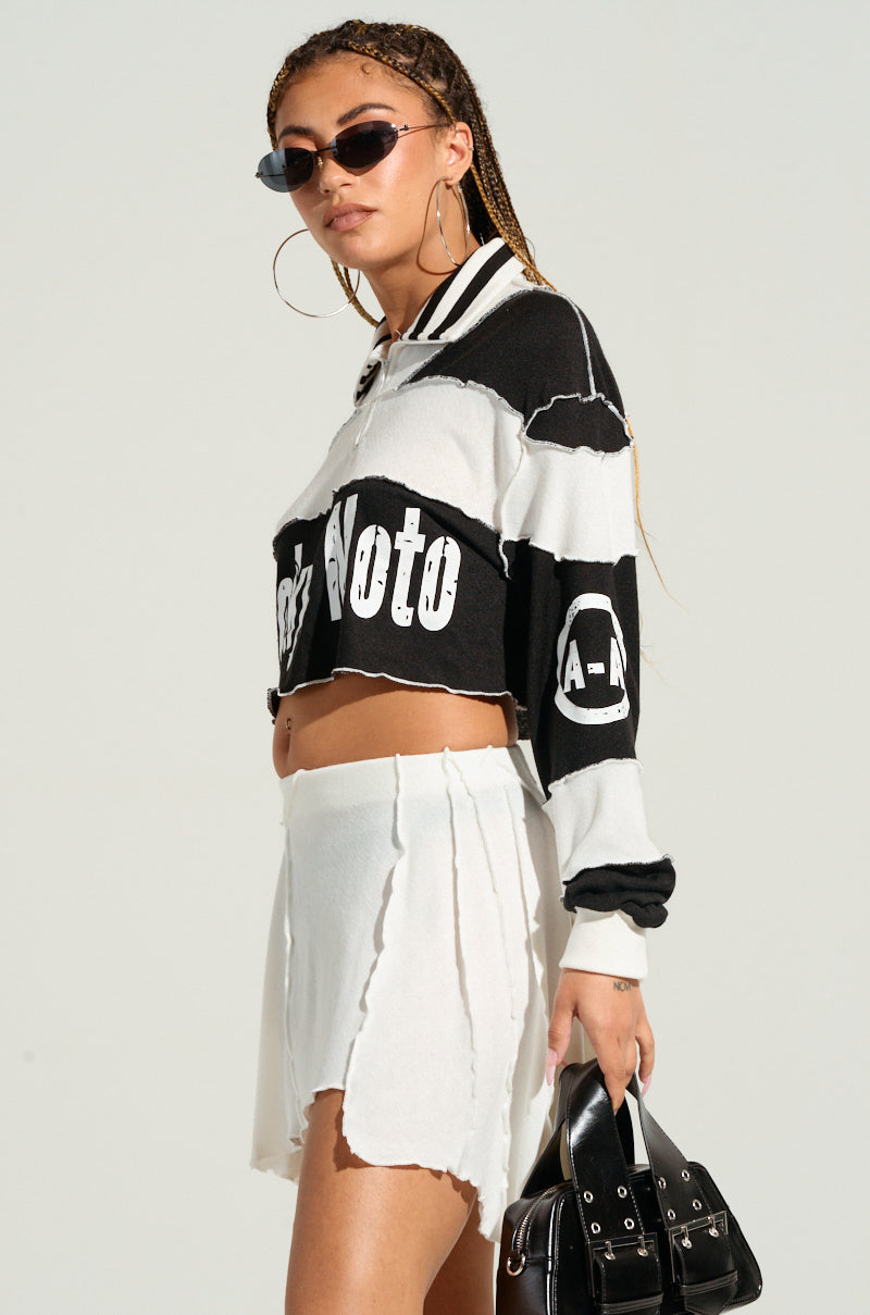 A woman with long, braided hair wearing sunglasses, a black and white "HANDMADE AND COOL TOP" featuring colorblock paneled construction, and a white skirt with ruffled edges. She is holding a black handbag. The outfit exudes a sporty, casual style reminiscent of TOKYO MOTO fashion.
