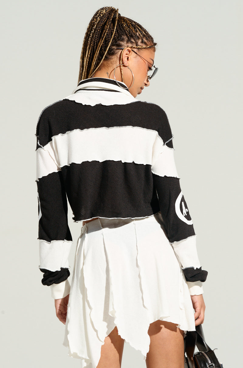 A person with braided hair is wearing a HANDMADE AND COOL TOP, featuring black and white stripes with colorblock paneled construction, paired with an asymmetrical white skirt. They are facing away from the camera, carrying a black bag against a plain light gray background.