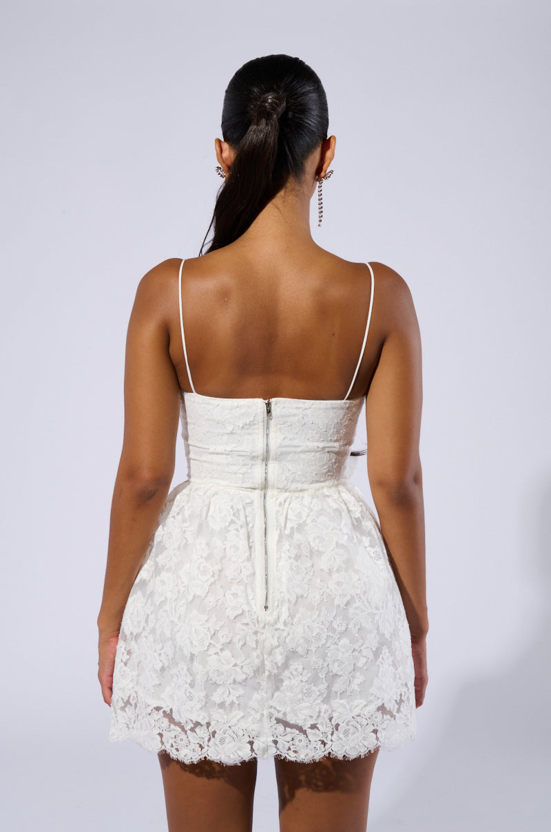 A woman is shown from the back, wearing the ARIANA LACE MINI DRESS, characterized by its white lace fabric, thin straps, and a visible zipper running down the center. Her hair is tied back in a sleek ponytail against a plain gray background.