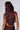 A person wearing the DESERT VIBE SLEEVELESS PU TOP IN BURGUNDY with their back to the camera showcases buckle embellishments. The hood covers their head, revealing tattoos on their left arm and shoulder. Matching burgundy pants are partially visible against a plain, gray background.