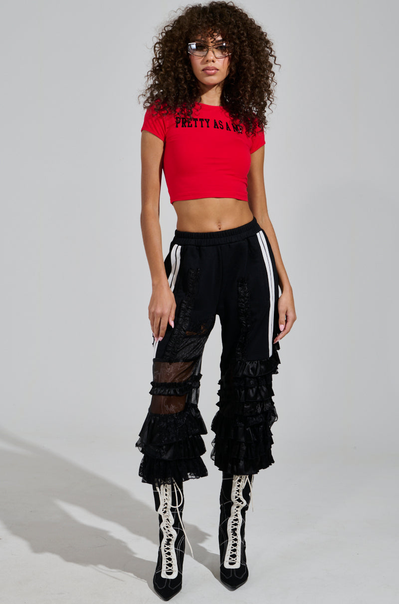 A person with curly hair wears sunglasses, a red cropped t-shirt, and the AKIRA Label's ALL OVER THE PLACE PANT, featuring ruffled mesh layers and an elastic waistband for comfort. They pair this outfit with white lace-up boots against a plain gray background.