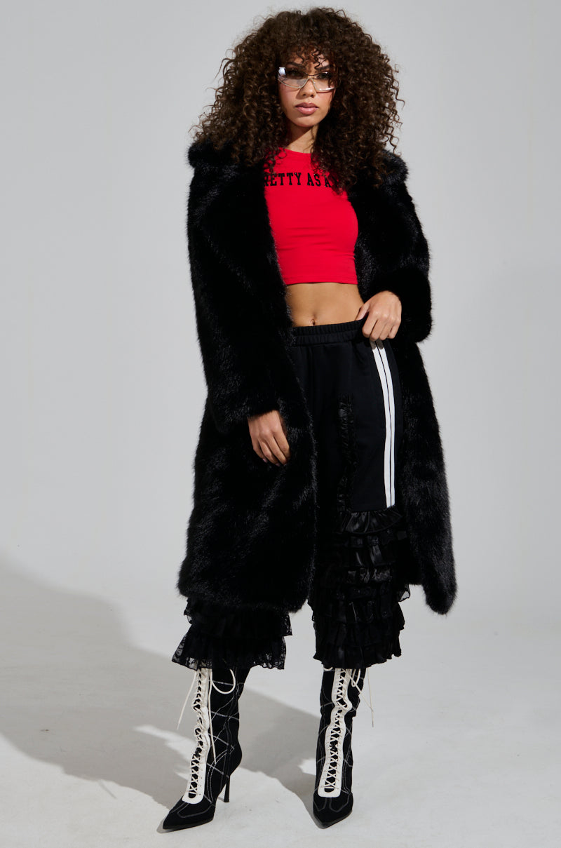 A person with curly hair wearing a red crop top and the "ALL OVER THE PLACE PANT" in black, featuring sheer mesh cutout panels, is paired with a black faux fur coat and lace-up heeled boots, posing against a plain background.