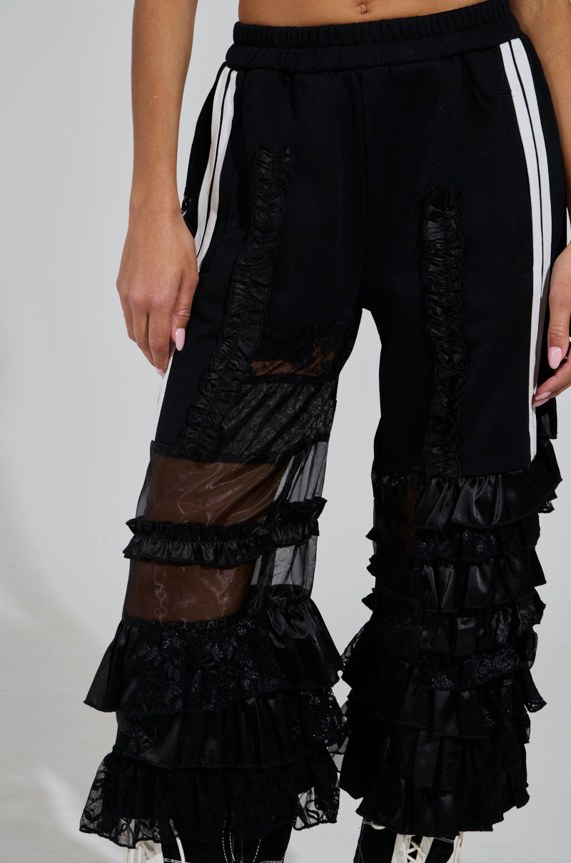 A person is wearing the ALL OVER THE PLACE PANT, characterized by its black color with white side stripes, high-rise design, and multiple layers of black ruffles and sheer fabric. The pants have a unique, textured appearance featuring sheer mesh cutout panels. The person's hand with painted nails is also visible.