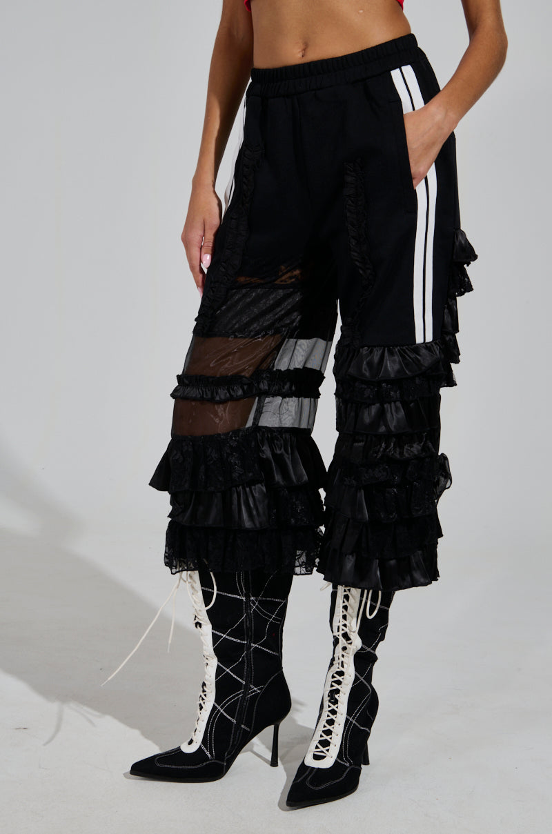 Against a plain background, the individual wears the ALL OVER THE PLACE PANT, featuring black ruffled layers and sheer mesh cutout panels with white side stripes. The look is finished off with black and white lace-up high-heeled boots.