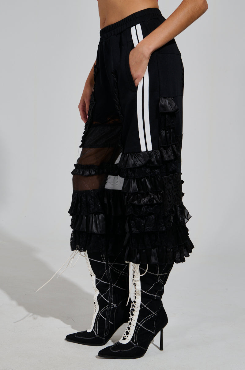 A person is wearing the ALL OVER THE PLACE PANT, which is black with white stripes and a high-rise design, featuring sheer mesh cutout panels for added texture. Complementing the outfit are black lace-up boots with standout white laces and high heels.