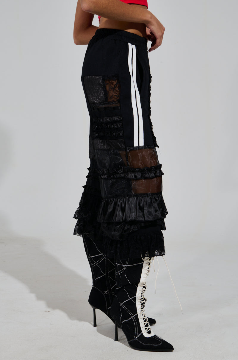 A person is wearing ALL OVER THE PLACE PANT in black with sheer mesh cutout panels, paired with black lace-up high-heeled boots featuring a geometric design. The outfit contrasts with a red top, visible in the background.