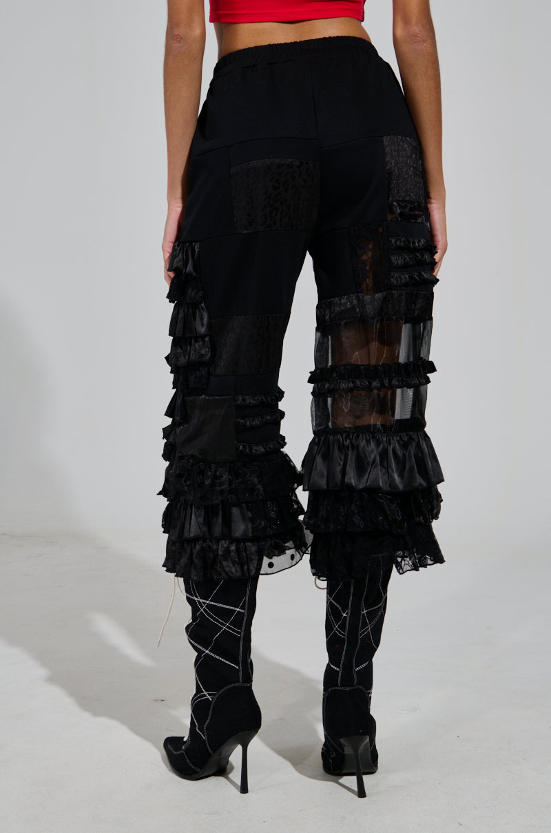 A person is wearing the ALL OVER THE PLACE PANT, which features high-rise styling, black sheer mesh cutout panels, and multiple ruffles on the legs. The ensemble is paired with high-heeled shoes that have a crisscross pattern. The background is a plain, neutral color.