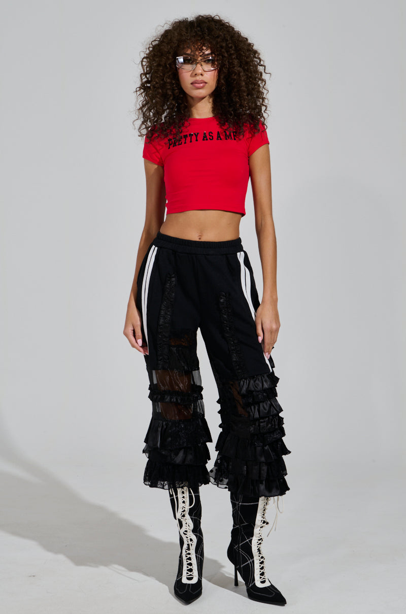 A person with curly hair is wearing a red crop top and black, ruffled high-rise ALL OVER THE PLACE PANT. They have on stylish glasses, knee-high black and white lace-up boots, with a hint of sheer mesh cutout panels adding an edgy flair.