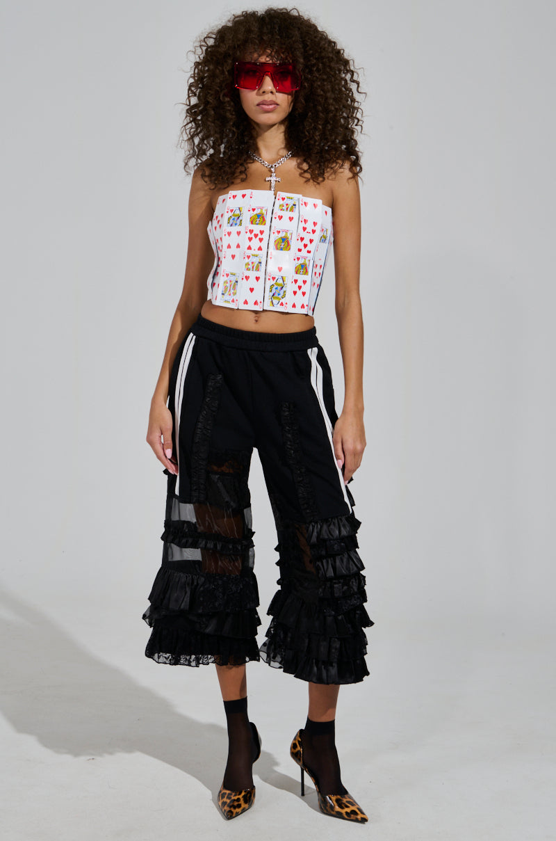 A person with curly hair and red sunglasses is wearing an AKIRA Label strapless top with card designs, the ALL OVER THE PLACE PANT featuring ruffled details and an elastic waistband, along with cheetah print shoes. They stand confidently against a plain backdrop.