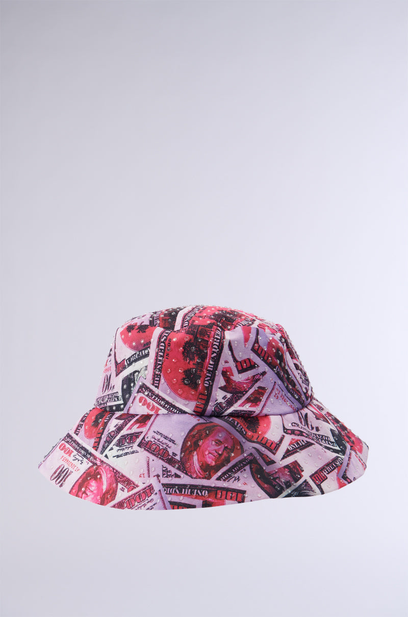 The MONEY ON MY MIND BUCKET HAT is a $100 collage-patterned bucket hat featuring red, pink, and white hues. It showcases various artistic denominations with rhinestone accents set against a plain light gray background.