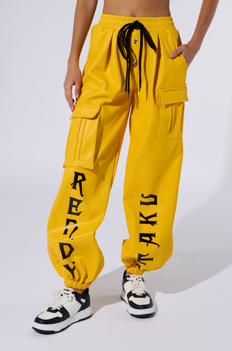 A person is wearing the DONE WITH YOU JOGGER, which are bright yellow high-rise cargo pants featuring "READY" on the left leg and "TAKIS" on the right. The pants have multiple cargo pockets and are paired with black and white sneakers. The background is plain white.