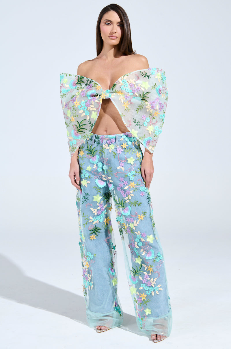 A woman confidently poses in GARDENIA DENIM WITH FLORAL TULLE WIDE LEG JEANS, featuring a sheer floral design with a large bow-like top. The colorful flowers on translucent fabric and a tulle overlay create an airy, stylish look against a plain, light background. 0