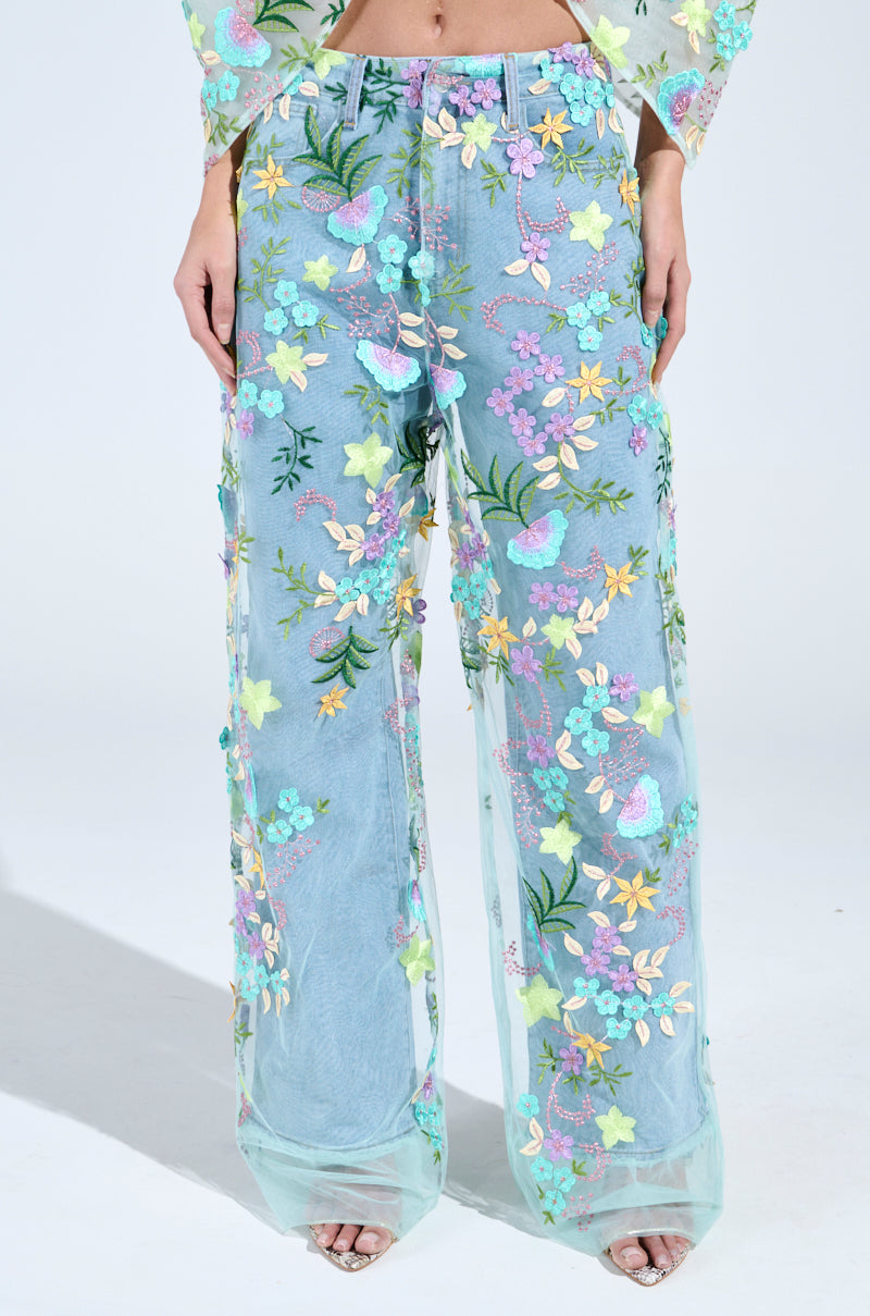 A person models the Gardenia Denim with Floral Tulle Wide Leg Jeans in light blue, featuring a sheer design adorned with colorful embroidered flowers and leaves, adding whimsy to its vibrant, transparent charm. 1