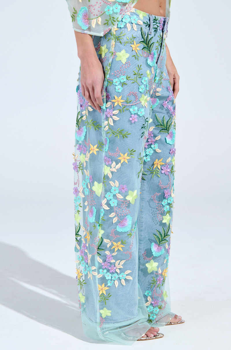 A person models GARDENIA DENIM WITH FLORAL TULLE WIDE LEG JEANS featuring pink, yellow, and green embroidery. The look is completed with open-toe heels against a white backdrop. 2