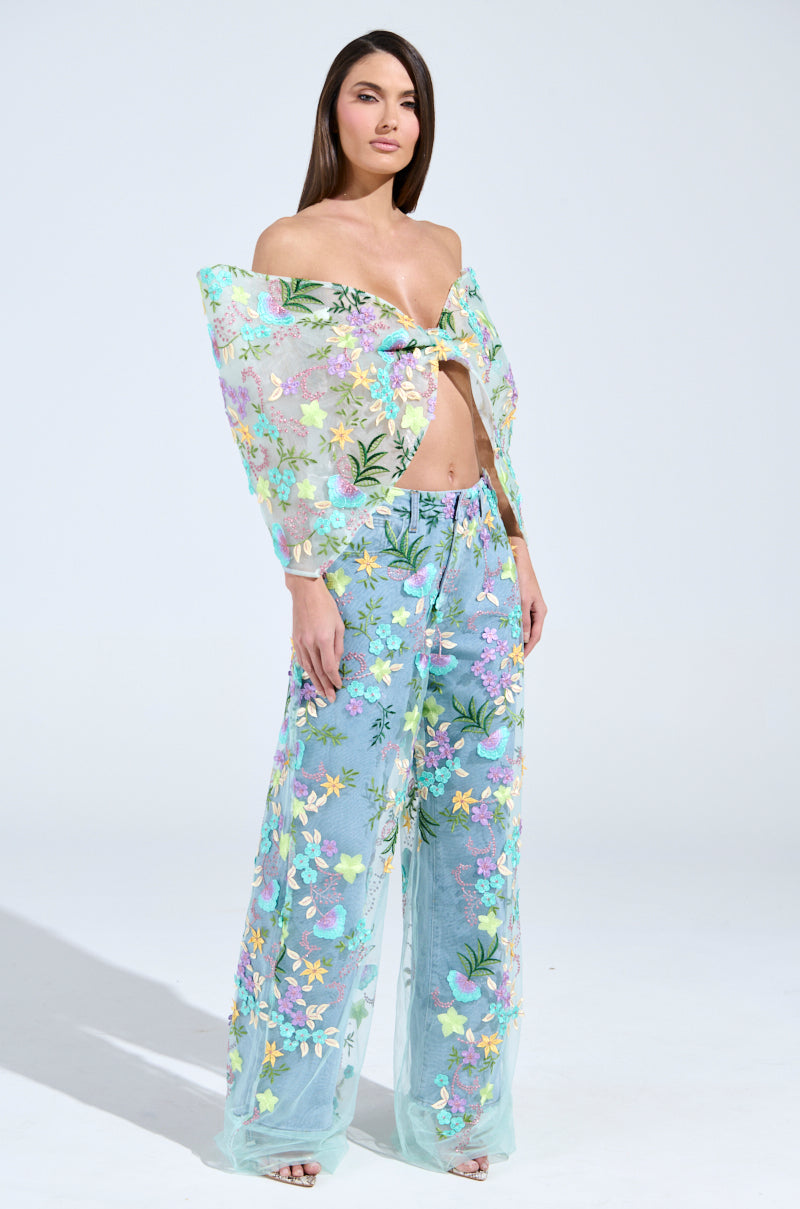 A woman wears the Gardenia jeans with a pastel sheer floral tulle overlay top and matching pants. Her off-the-shoulder look with a prominent front bow stands out against a light background. 4